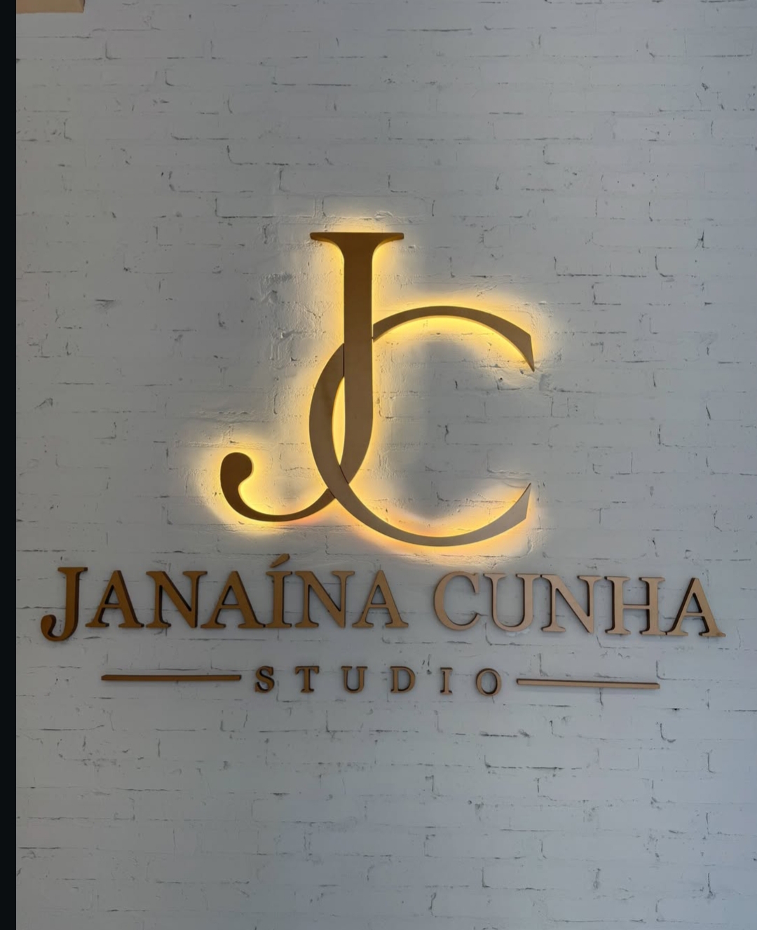 Letreiro dourado iluminado do Janaína Cunha Studio na parede de tijolos brancos